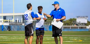 A coach holding notes speaks with two football players on a field.