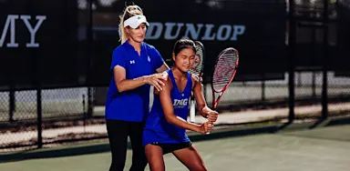 An IMG Academy coach guides a student-athlete tennis player’s stance and grip during practice on an outdoor court, adjusting her positioning as she prepares for a shot.