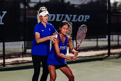 An IMG Academy coach guides a student-athlete tennis player’s stance and grip during practice on an outdoor court, adjusting her positioning as she prepares for a shot.