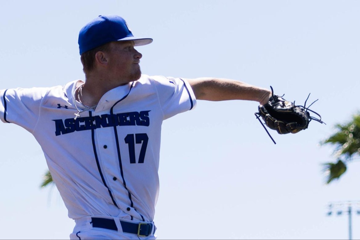 Baseball pitcher in mid-throw during a game, wearing a white "Ascenders" jersey with palm trees in the background.