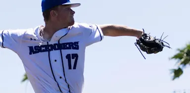 Baseball pitcher in mid-throw during a game, wearing a white "Ascenders" jersey with palm trees in the background.