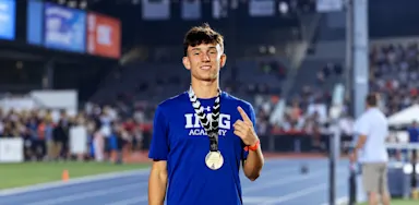 An athlete wearing medals smiles and raises one finger at a sports venue.