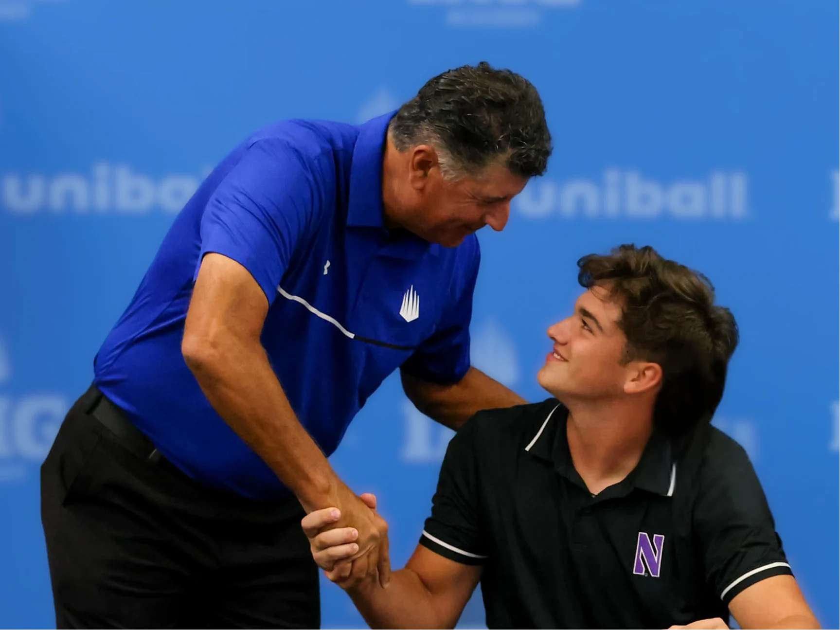 An IMG Academy coach shakes hands with a student-athlete during a signing moment, both smiling as they make eye contact in front of a branded backdrop.
