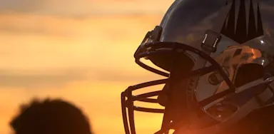 Football player in reflective helmet with black logo silhouetted against a sunset sky.