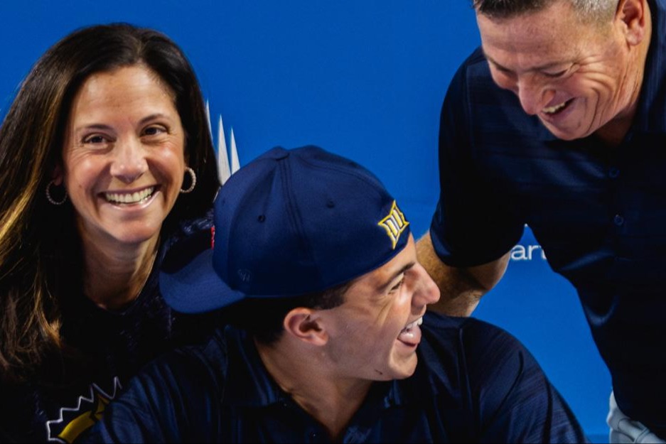 A smiling young man in a sports cap sits between his parents as they all laugh and celebrate in front of a blue IMG Academy backdrop, suggesting a signing or commitment event.