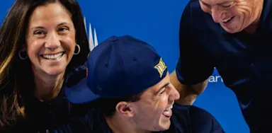 A smiling young man in a sports cap sits between his parents as they all laugh and celebrate in front of a blue IMG Academy backdrop, suggesting a signing or commitment event.