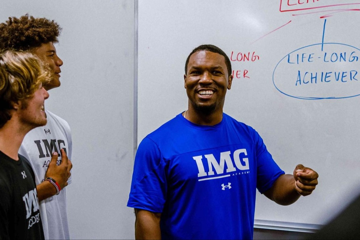 Coach leading a group of student-athletes in a classroom at IMG Academy, standing in front of a whiteboard with the phrase "Life-Long Achiever" written on it.