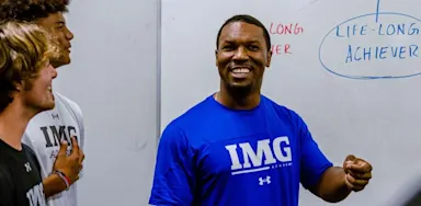 Coach leading a group of student-athletes in a classroom at IMG Academy, standing in front of a whiteboard with the phrase "Life-Long Achiever" written on it.