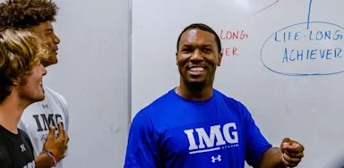 A smiling coach speaks beside a whiteboard in a classroom.