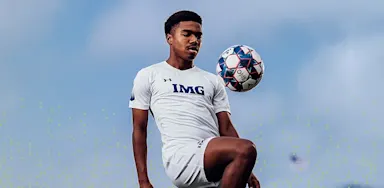 IMG Academy soccer athlete in white training kit controlling a soccer ball mid-air during practice, demonstrating balance and technical skill against a blue sky.