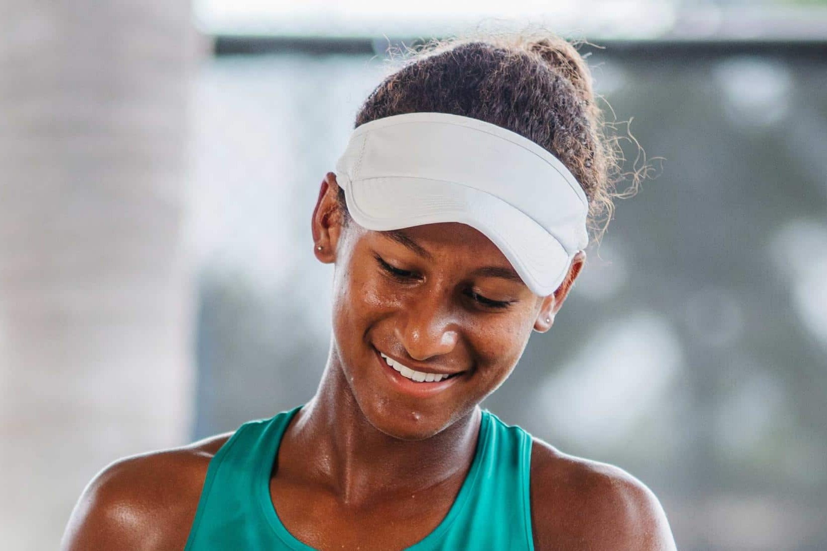 Young female tennis player smiling after a training session, wearing a white visor and teal athletic top.
