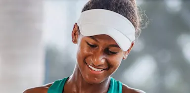 Young female tennis player smiling after a training session, wearing a white visor and teal athletic top.