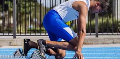 Male IMG Academy track athlete kneels at the starting line on a bright blue track, preparing for a sprint and focusing on the start of the race or practice.