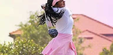 An IMG Academy student-athlete golfer follows through on a swing, wearing a pink visor and skirt, with a building and greenery in the background.