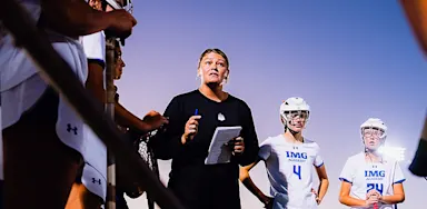 A coach holding a clipboard addresses a group of lacrosse players on an outdoor field.