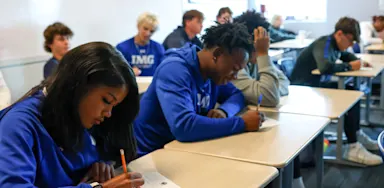 IMG Academy students sit at desks in a bright classroom, focused on writing during a sports marketing or academic exercise, wearing branded blue athletic apparel.