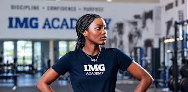 An athlete stands with her hands on her hips in a weight room.