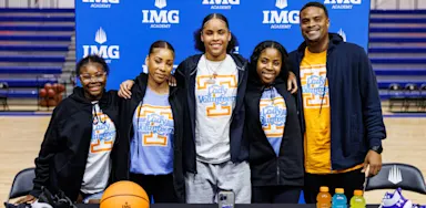 IMG Academy basketball player stands with family on signing day, wearing a Tennessee shirt behind a table with a ball, shoes, and drinks, celebrating her college commitment.