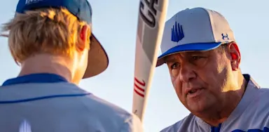 An IMG Academy baseball coach speaks intently to a young athlete during practice, holding a bat vertically as they discuss technique as the sun sets in the background.