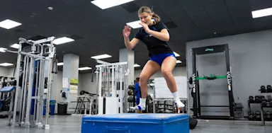 An athlete performs a jump onto a padded box in a gym.