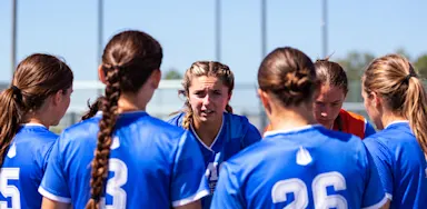 A soccer player speaks to teammates gathered in a huddle on the field.