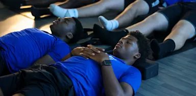 Student-athletes in blue shirts focusing on deep breathing and recovery after a workout, emphasizing rest and sleep for muscle repair.