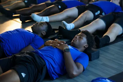 Student-athletes in blue shirts focusing on deep breathing and recovery after a workout, emphasizing rest and sleep for muscle repair.