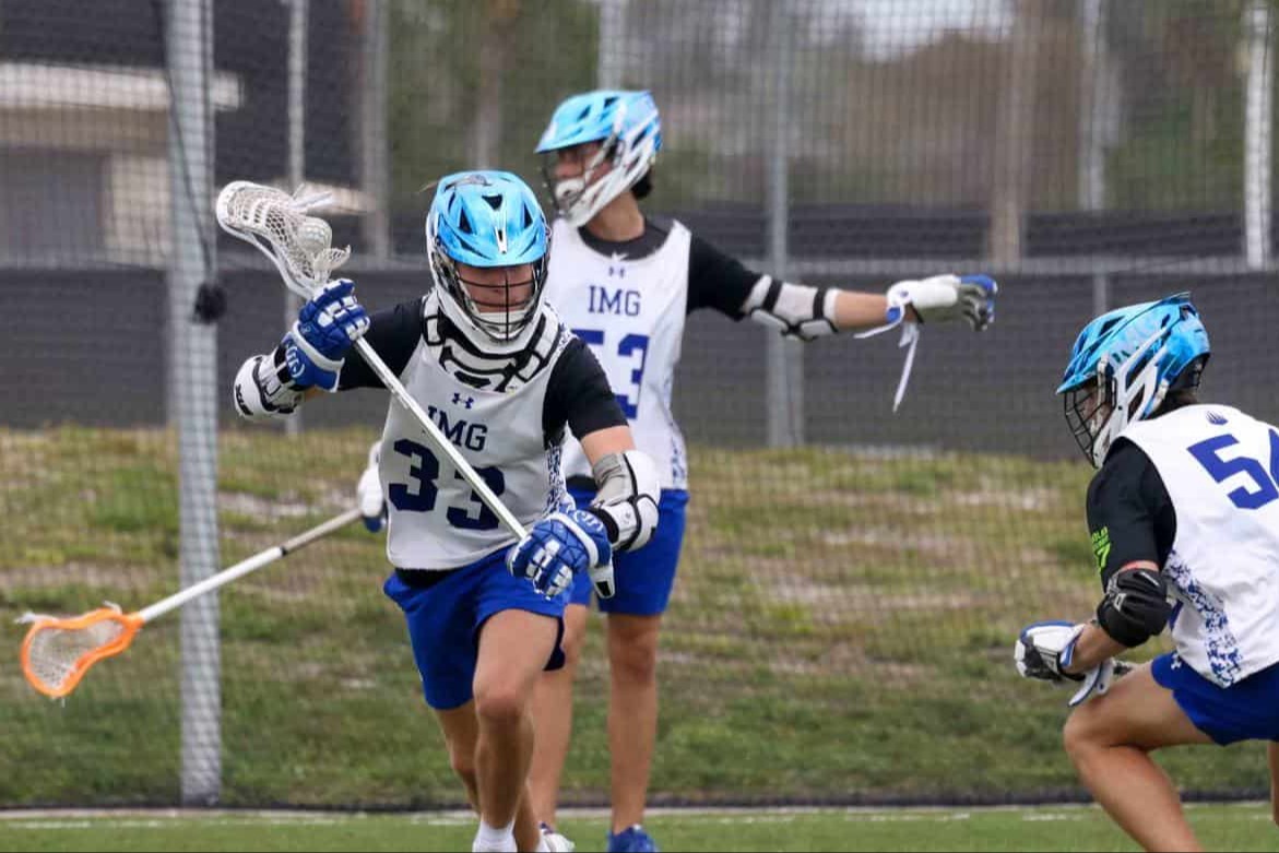 Three IMG Academy lacrosse players in helmets and practice gear actively scrimmaging on a field.