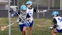 Three IMG Academy lacrosse players in helmets and practice gear actively scrimmaging on a field.
