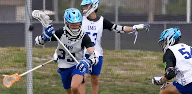 Three IMG Academy lacrosse players in helmets and practice gear actively scrimmaging on a field.