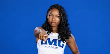 An athlete in a white team jersey points toward the camera against a blue background.
