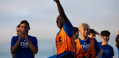 Boys Soccer athlete in IMG Academy gear cheering loudly and pointing skyward during a competitive practice session or game day huddle.