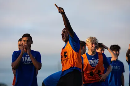 Boys Soccer athlete in IMG Academy gear cheering loudly and pointing skyward during a competitive practice session or game day huddle.