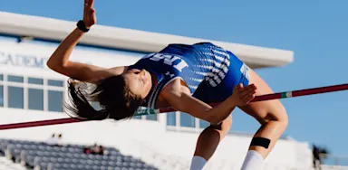 An IMG Academy student-athlete high jumper clears the bar with an arched back during a track and field event, with stadium seating in the background.