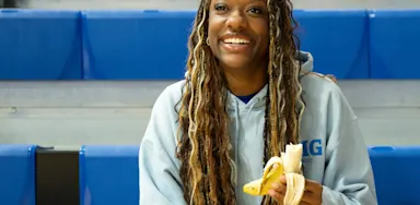 IMG Academy student-athlete sits on blue bleachers smiling, wearing a light hoodie and holding a partially peeled banana, suggesting a healthy snack during practice or downtime.