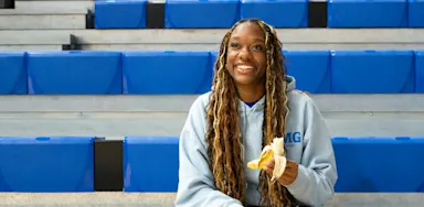 An athlete smiles while holding a peeled banana on gym bleachers.