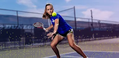 Female tennis athlete wearing an IMG Academy uniform hits a forehand on an outdoor tennis court, focused on the ball during training under a clear blue sky.