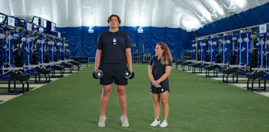 An athlete holds dumbbells at his sides during a calf-raise exercise beside a trainer.