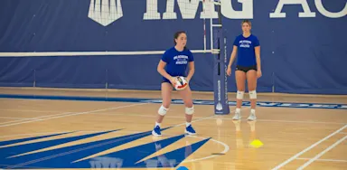 Two volleyball players stand beside cones for a setting drill.