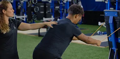 A trainer guides an athlete performing a resistance-band exercise attached to a rack.