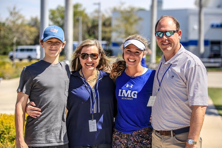 A smiling family of four poses outside on check in day at IMG Academy on a sunny day. The young woman in the center wears an IMG Academy athletic shirt and visor, standing next to her parents and brother.