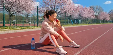 A student-athlete sits on a running track eating a snack beside a water bottle.