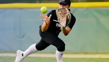 A softball player from IMG Academy fields a ground ball, crouching low with her glove extended and eyes focused on the ball as it approaches. She is wearing a black uniform, white knee-high socks, and a black visor, positioned on a grass field with an outfield fence in the background.