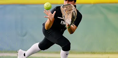A softball player from IMG Academy fields a ground ball, crouching low with her glove extended and eyes focused on the ball as it approaches. She is wearing a black uniform, white knee-high socks, and a black visor, positioned on a grass field with an outfield fence in the background.