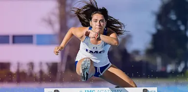 IMG Academy track and field athlete clears a hurdle during a sprint hurdle event, captured midair with strong form and focus on an outdoor track.