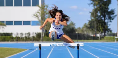 A track athlete clears a hurdle on a blue running track.