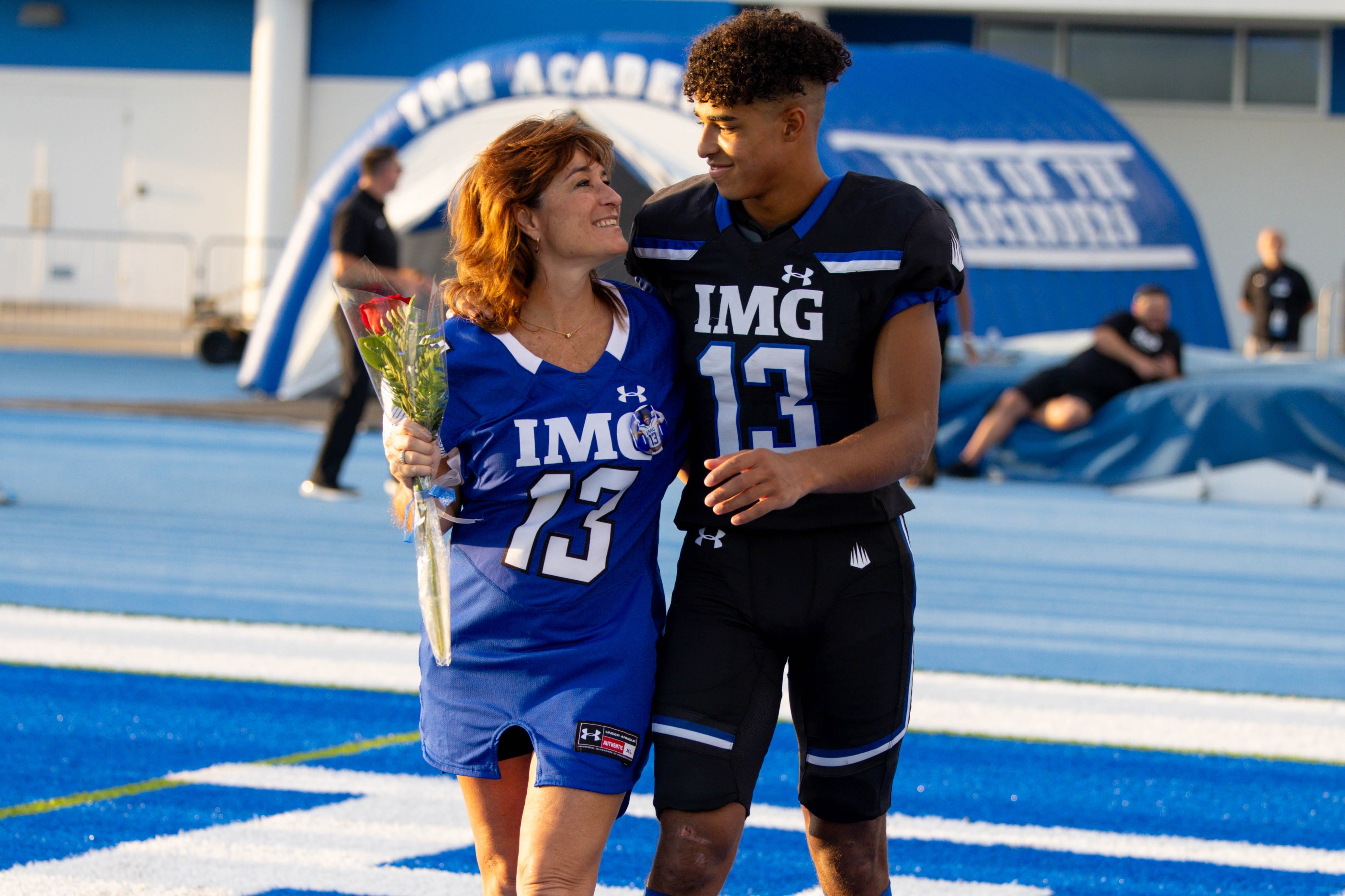 Youth football player in a black and blue IMG Academy uniform standing with his mother on the blue turf during Senior Night or Parent's Weekend.
