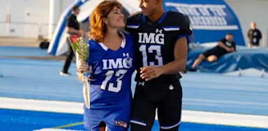 Youth football player in a black and blue IMG Academy uniform standing with his mother on the blue turf during Senior Night or Parent's Weekend.