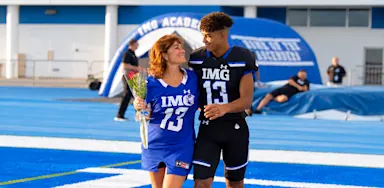 Youth football player in a black and blue IMG Academy uniform standing with his mother on the blue turf during Senior Night or Parent's Weekend.