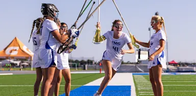 Female lacrosse players celebrating the start of a game on the IMG turf field, illustrating team spirit and athletic excellence in women's sports.